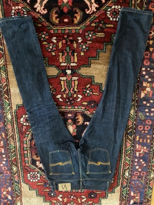Mörkblå slim jeans från Nudie Jeans - Snygga mörkblå slim jeans från Nudie Jeans med klassiska orange sömmar och broderade bakfickor. Jeansen har låg midja och smal passform, tillverkade i slitstark denim. Perfekta för dig som gillar en stilren och modern look. I storleken 28W och 32L