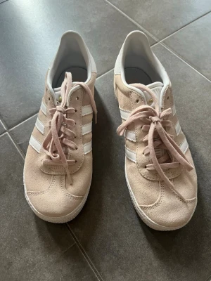 Ljusrosa Adidas sneakers med mocka, stl 38 - Snygga ljusrosa sneakers från Adidas i mocka med klassiska vita ränder på sidorna och vita detaljer. Skorna har lågt skaft och platt sula med mönster undertill. Perfekta för en stilren och avslappnad look. Jättefint skick, knappt använda, i princip som nya! 