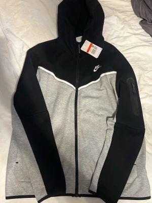 Nike Tech Fleece hoodie svart/grå - Nike Tech Fleece hoodie i svart och grått med dragkedja framtill, vit piping och ficka med dragkedja på ärmen. Klassisk Nike-logga på bröstet och huva. Tillverkad i mjukt och lätt material som passar perfekt till streetwear-stilen.