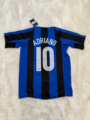 Inter Adriano #10 Nike fotbollströja - Säljer en klassisk Inter Milan fotbollströja med Adriano och nummer 10 på ryggen. Tröjan är blå och svart med breda ränder, korta ärmar och Pirelli som sponsor på bröstet. Officiell Nike-logga och Inter-emblem på framsidan. Perfekt för dig som älskar retro och italiensk fotboll.