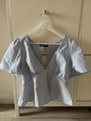 Randig blus med v-ringning  - Snygg blus från med blåvita ränder, v-ringning och kort ärm. Toppen har knappar framtill, lätt puffiga ärmar och en öppen detalj i ryggen med knyt. Perfekt för dig som gillar trendiga och luftiga toppar, perfekt till sommaren och våren💕