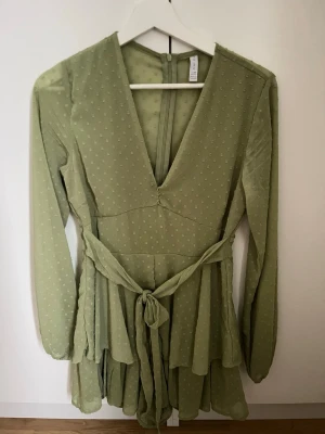 Ljusgrön prickig omlottklänning H&M - Säljer en ljusgrön kortklänning från H&M med prickigt mönster, v-ringning och omlottdetalj i midjan. Klänningen har långa ärmar, volangkant nertill och knytband i midjan. Materialet är lätt och luftigt, perfekt för varmare dagar.