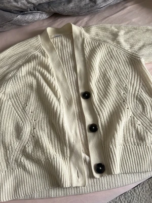 Vit stickad kofta med svarta knappar - Snygg vit stickad kofta med tre stora svarta knappar framtill. Koftan har ett grovt stickat mönster och bred ribbad kant nedtill och vid ärmslut. Perfekt att slänga över en t-shirt för en chill look. Passar bra för mig som har storlek S