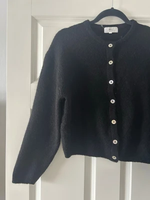 Svart ullkofta med pärlemorknappar - Mysig svart kofta med rund halsringning och pärlemorskimrande knappar framtill. Koftan har lång ärm och är stickad i ett mjukt material som känns skönt mot huden. Perfekt att slänga på sig när det är lite kyligt ute.