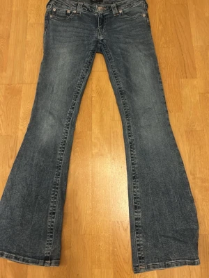 Blå bootcut jeans från true religion  - Ett par klassiska blå bootcut jeans från true religion med lätt slitna detaljer och fem fickor. Jeansen har låg midja och vida ben, perfekt för en avslappnad och trendig look. Tillverkade i denim med snygga sömmar och knappgylf framtill.