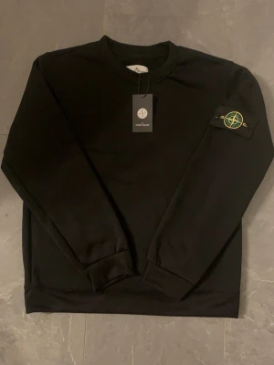 Svart sweatshirt från Stone Island - Snygg svart sweatshirt från Stone Island med klassisk rund hals och ikonisk logotyp-patch på vänster ärm. Tröjan har ribbade muddar vid ärmslut och nederkant. Perfekt för en clean och stilren look.