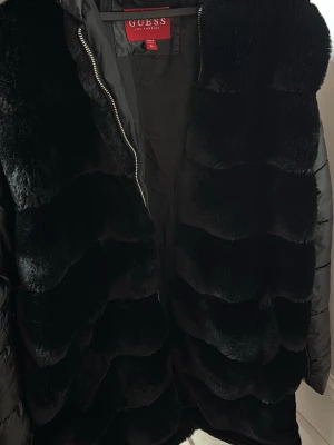 Svart pälsjacka från Guess - Snygg svart pälsjacka från Guess med glansig fuskpäls framtill och quiltade ärmar. Jackan har dragkedja och en modern, oversized look. Perfekt för dig som vill ha en trendig vinterjacka med lyxig känsla.