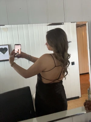 Svart satinklänning med tunna band - Säljer en svart satinklänning med tunna axelband och snygg draperad urringning. Klänningen har en slimmad passform och ett glansigt material som ger en lyxig känsla. Öppenrygg med dekorativa banddetaljer som gör den extra speciell.