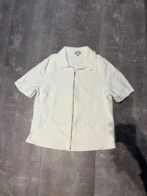 Vit kortärmad skjorta från COS - Vit skjorta från COS med korta ärmar och klassisk krage. Skjortan har dragkedja framtill och är tillverkad i ett ribbat material som ger en modern känsla. Perfekt för en stilren och avslappnad look.