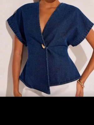 Mörkblå omlottopp i denim - Snygg mörkblå omlottopp i denim med korta vida ärmar och markerad midja. Toppen har en unik knappdetalj framtill och en djup v-ringning. Perfekt för dig som vill sticka ut med en cool och trendig look.