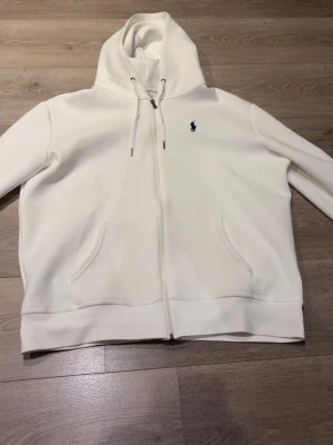Vit hoodie från Polo Ralph Lauren, ny skick - Snygg vit hoodie från Polo Ralph Lauren med dragkedja framtill och två fickor. Klassisk huva med dragsko och den ikoniska lilla loggan broderad på bröstet. Tillverkad i mjuk bomull som känns riktigt skön mot huden. Perfekt för en clean och stilren look. Ny skick aldrig använd mvh 