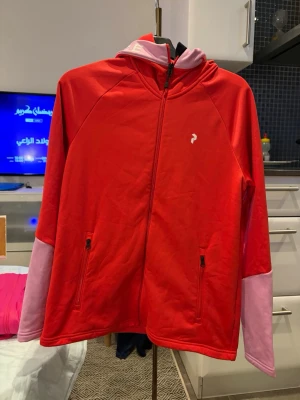 Röd hoodie från Peak Performance - Snygg röd hoodie från Peak Performance med dragkedja och två fickor framtill. Huva med ljusrosa insida och textdetalj. Ärmarna har rosa partier vid handlederna. Perfekt för en sportig och avslappnad stil.