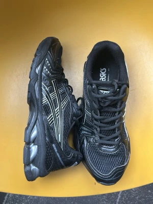 Asics gel kayano 14 black colorway - Svarta sneakers från ASICS med silvriga detaljer och meshpaneler för bra ventilation. Skorna har en chunky sula i svart och grått, samt klassiska ASICS-loggor på sidorna. Perfekta för dig som gillar sportig och retro stil.