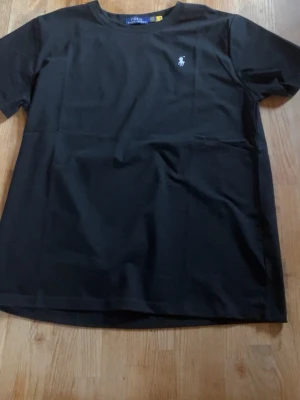 Svart t-shirt från Ralph Lauren - Klassisk svart t-shirt från Ralph Lauren med den ikoniska broderade loggan på bröstet. T-shirten har rund halsringning och korta ärmar, perfekt för en stilren och enkel look.