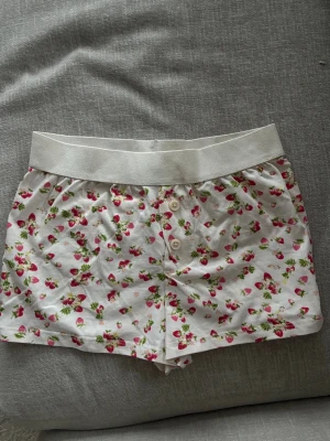 Vita pyjamasshorts med jordgubbsmönster - Söta pyjamasshorts i vitt med små röda jordgubbar och gröna blad. Shortsen har bred resår i midjan och två dekorativa knappar framtill. Perfekta för mysiga hemmakvällar och tillverkade i mjukt bomullstyg.