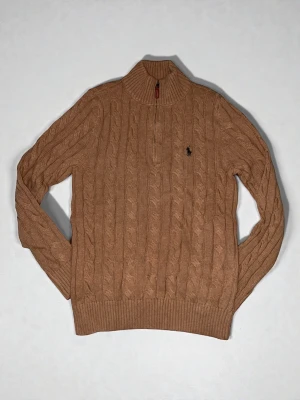 Beige kabelstickad tröja Polo Ralph Lauren - Säljer en beige kabelstickad tröja från Polo Ralph Lauren med halv dragkedja och hög krage. Tröjan har det klassiska broderade logotypen på bröstet och ribbade muddar vid ärmslut och nederkant. Perfekt för dig som gillar stilren och tidlös design.
