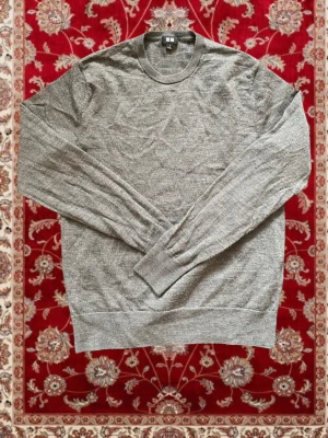 Grå stickad tröja från Uniqlo - En stilren grå stickad tröja från Uniqlo med rund halsringning och långa ärmar. Tröjan har ribbade muddar vid ärmslut och nederkant. Perfekt basplagg i mjukt material som passar till allt. Obs ett håll vid armvecket!!!