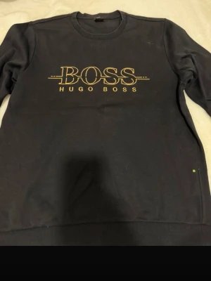 Svart sweatshirt från Hugo Boss - Snygg svart sweatshirt från Hugo Boss med gul logga och text på bröstet. Klassisk rund halsringning och ribbade muddar vid ärmar och nederkant. Perfekt för en avslappnad och trendig stil. Storlek L passar på M 
