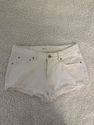 Vita jeansshorts från Gina Tricot - Säljer ett par vita jeansshorts . De har råa, fransiga kanter och klassisk femficksdesign med knapp och dragkedja framtill. Perfekta för varma dagar och enkla att matcha med olika toppar.