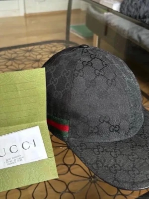 Gucci keps svart - Snygg svart keps från Gucci med klassiskt GG-monogram över hela kepsen. Kepsen har en böjd skärm och detaljer i grönt och rött på sidan. Tillverkad i canvasmaterial som ger en exklusiv känsla. Perfekt för dig som gillar streetstyle och lyxiga accessoarer.