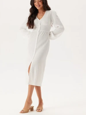 Vit ribbad långklänning med slits - Vit långklänning med ribbad struktur och v-ringning. Klänningen har långa ärmar, knappar framtill och en snygg slits. Perfekt för dig som ska ta studenten!