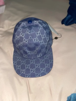 Blå Gucci keps med monogram - Snygg blå keps från Gucci med klassiskt GG-monogram över hela kepsen. Kepsen har böjd skärm och är tillverkad i ett denimliknande material. Perfekt accessoar för att lyfta din outfit med en lyxig touch.