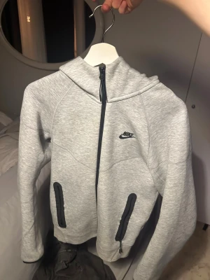 Grå nike tech generation 4 XS - Snygg grå hoodie från Nike i storlek XS. Hoodien har hel dragkedja, huva och svarta detaljer vid fickorna. Perfekt för chill eller träning, med klassisk Nike-logga på bröstet. Mjuk och skön i bomullsmix, långärmad och med modern passform. Den är äkta köpte den ifrån zalando för 1400kr