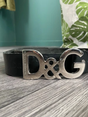 Svart D&G bälte med silverdetalj - Snyggt svart bälte från Dolce & Gabbana med stor silverfärgad D&G-logga som spänne. Bältet är i skinn och har präglade D&G-loggor längs hela remmen. Perfekt för att lyfta vilken outfit som helst.