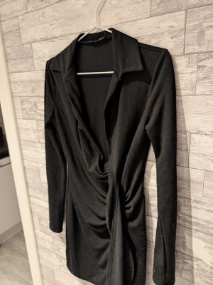 Svart omlottklänning med knyt - Stilren svart omlottklänning med lång ärm och snygg knytning i midjan. Klänningen har en djup v-ringning och krage, vilket ger en trendig look. Perfekt för dig som gillar minimalistisk stil och vill ha något enkelt men ändå elegant.