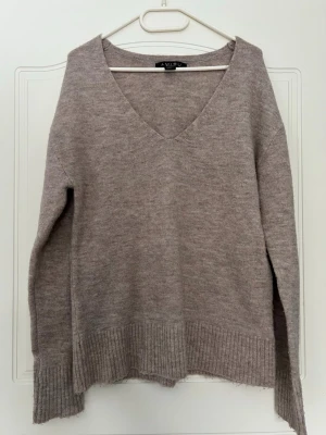 Beige/brun stickad tröja med v-ringning - Mysig beige/brun stickad tröja från amisu med djup v-ringning och långa ärmar. Tröjan har ribbstickade muddar vid ärmslut och nederkant, samt en avslappnad passform som gör den perfekt för lager på lager. Tillverkad i mjukt material. Stor i storleken! Perfekt att gå runt med hemma! 
