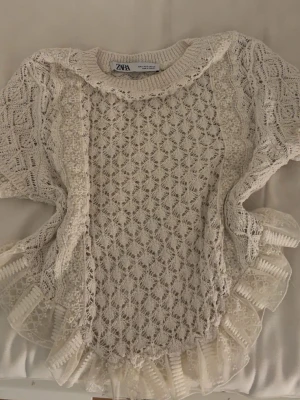 Vit spetsblus med volang från Zara - Supersöt vit spetsblus från Zara med korta ärmar och volangdetaljer längs kanterna. Blusen har ett genombrutet spetsmönster och dekorativa volanger i nederkant och längs sidorna. Perfekt för dig som gillar romantisk och feminin stil.