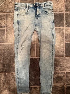 Ljusblå slimfit Replay anbass  - Säljer ett par ljusblå slim fit jeans från Replay med klassisk femficksdesign och diskret slitning på vänster ben. Jeansen har en snygg tvättad look och tydliga sömmar längs benen. Perfekta för en avslappnad och trendig stil. Ett litet hål på knät