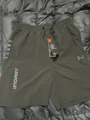 Svarta shorts från Under Armour - Säljer ett par svarta shorts från Under Armour med färgglada detaljer och logga på vänster ben. Shortsen har elastisk midja med snörning och är tillverkade i ett lätt och bekvämt material, perfekta för träning eller chill.