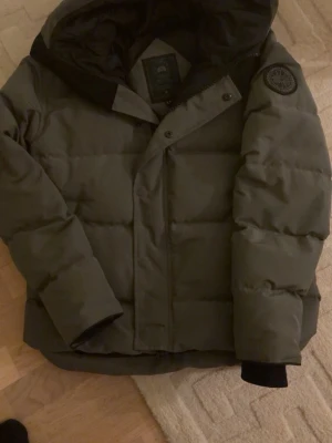 Mörkgrå pufferjacka från Canada Goose - Snygg mörkgrå pufferjacka från Canada Goose med huva och diskret logotyp på ärmen. Jackan har dragkedja och tryckknappar framtill samt ribbade muddar vid ärmsluten. Perfekt för kalla dagar och riktigt varm tack vare det tjocka fodret.