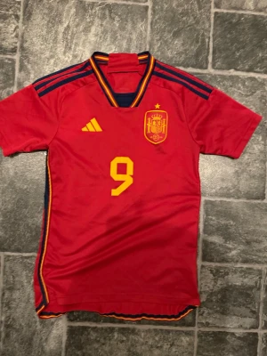 Spaniens landslagströja Gavi #9 - Röd matchtröja från Spaniens landslag med gula detaljer, nummer 9 och namnet Gavi på ryggen. Tröjan har korta ärmar, blå ränder på axlarna och broderat landslagsmärke på bröstet. Tillverkad i lätt och ventilerande material, perfekt för fotboll. ÄKTA FRÅN UNISPORT 