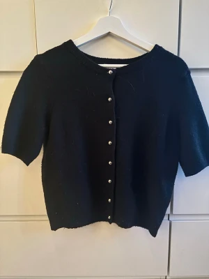 Svart kortärmad kofta med pärlknappar - Stilren svart kofta med korta ärmar och rund halsringning. Koftan har dekorativa pärlknappar framtill och är gjord i ett mjukt, stickat material. Perfekt att slänga över en topp för en enkel men chic look. Nästan aldrig använd 