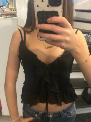 Svart volangig linnetopp från Amisu\Newyorker  - Supersöt svart linnetopp från Amisu med tunna axelband, volangdetaljer och lätt transparent tyg. Toppen har en croppad passform och v-ringad urringning, perfekt att styla med jeans eller kjol. Materialet känns lätt och luftigt.