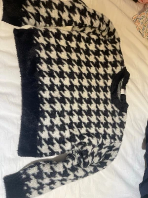Stickad tröja med hundtandsmönster - Säljer en fluffig stickad tröja i svart och vitt med klassiskt hundtandsmönster. Tröjan har rund halsringning, långa ärmar och ribbade muddar. Perfekt för dig som gillar retrovibbar och vill ha något riktigt mysigt i garderoben.