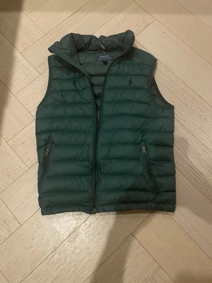 Mörkgrön dunväst från Polo Ralph Lauren - Snygg mörkgrön dunväst från Polo Ralph Lauren med quiltad design och diskret logga på bröstet. Västen har hög krage, dragkedja framtill och två fickor med dragkedja. Perfekt att slänga över en hoodie eller tröja för extra värme. Storlek xs Den har en liten defekt på insidan som dock är lagad inget som påverkar funktionen då man ej ser den
