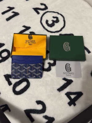 Blå korthållare från Goyard - Snygg blå korthållare från Goyard med klassiskt mönster i vitt och grått. Tillverkad i canvas och läder, perfekt för kort och ID. Kommer med originalask, dustbag och äkthetskort. En stilren accessoar för dig som gillar exklusiva detaljer.