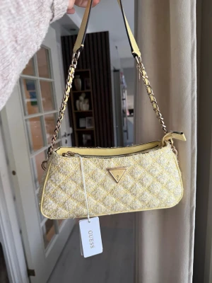 Gul quiltad handväska från Guess - Trendig gul handväska från Guess i quiltad tweed med kedjedetaljer på axelremmen och dragkedja upptill. Väskan har en avlång form och Guess-logga i metall framtill. Perfekt accessoar för att liva upp din outfit. Hör av dig vid intresse.