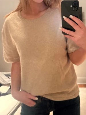 Beige stickad t-shirt med kort ärm - Säljer en enkel och stilren beige stickad t-shirt med korta ärmar och rund hals. Tröjan har ribbade muddar vid ärmslut och nederkant. Perfekt att matcha med jeans eller kjol för en avslappnad look.