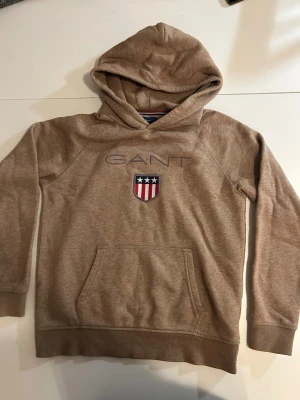 Beige hoodie från GANT med logga - Snygg beige hoodie från GANT med broderad logga och sköld på bröstet. Klassisk känguruficka framtill och huva. Mjuk bomullsblandning och ribbade muddar vid ärmslut och nederkant. Perfekt för en chill och stilren look.