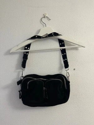 Svart handväska  - Cool svart handväska med bred axelrem. Väskan har två framfickor med dragkedjor och en större huvudficka. Perfekt för dig som gillar en edgy stil och vill ha plats för det viktigaste.