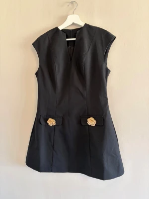 Svart kort klänning med gulddetaljer - Snygg svart kort klänning med v-ringning och ärmlös design. Klänningen har två dekorativa guldfärgade blommor framtill och markerade axlar för en cool siluett. Perfekt för dig som vill ha en stilren men unik look.