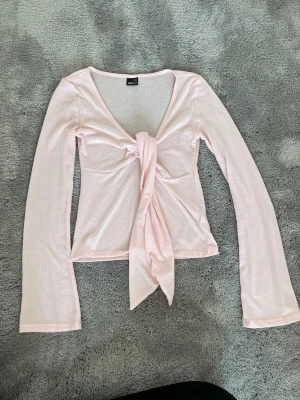 Ljusrosa knytblus från Gina Tricot - Supersöt ljusrosa blus från Gina Tricot med lång ärm och djup v-ringning. Toppen har en snygg knytning framtill som ger en trendig look. Tillverkad i mjuk bomullsmix som känns skön mot huden. Perfekt för att lyfta vilken outfit som helst!