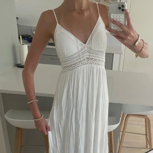 Vit långklänning med spetsdetaljer - Superfin vit långklänning med smala axelband och v-ringning. Klänningen har vackra virkade spetsdetaljer under bysten och vid urringningen, samt en flowy kjol i lätt, skrynkligt tyg. Perfekt för sommarens varma dagar och har en romantisk vibe.