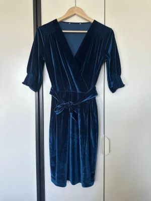 Marinblå omlottklänning i sammet - Snygg marinblå omlottklänning i mjuk sammet med trekvartsärm och knytband i midjan. Klänningen har v-ringad hals och smockdetaljer vid axlar och ärmslut för extra stilpoäng. Perfekt för dig som vill ha en elegant look med skön känsla.