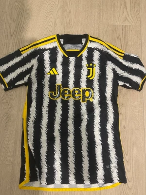 Juventus matchtröja svartvit Adidas - Juventus officiella matchtröja från Adidas med svartvita borstade ränder och gula detaljer. Tröjan har klubbmärke och tre stjärnor på bröstet, Jeep-logga i gult samt gula ränder på axlar och krage. Tillverkad i lätt och ventilerande material, perfekt för fotboll.