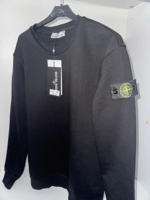 Svart sweatshirt från Stone Island - Säljer en svart sweatshirt från Stone Island med den ikoniska logotypen på vänster ärm. Tröjan har rund halsringning och ribbade muddar vid ärmslut och nederkant. Perfekt för en clean och stilren look. Storlek M men passar också för S
